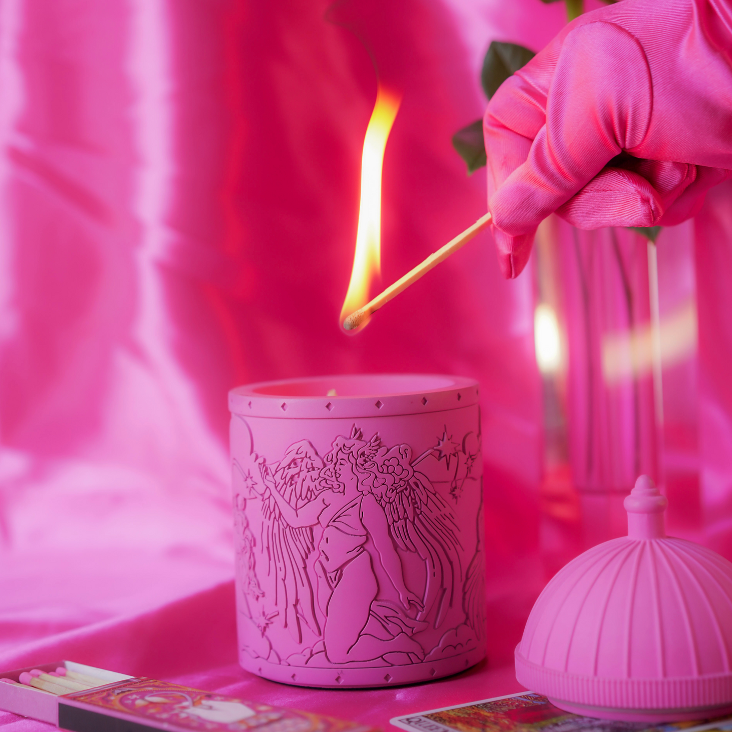 Pink candle with intricate designs on a pink background, with a match being lit.
